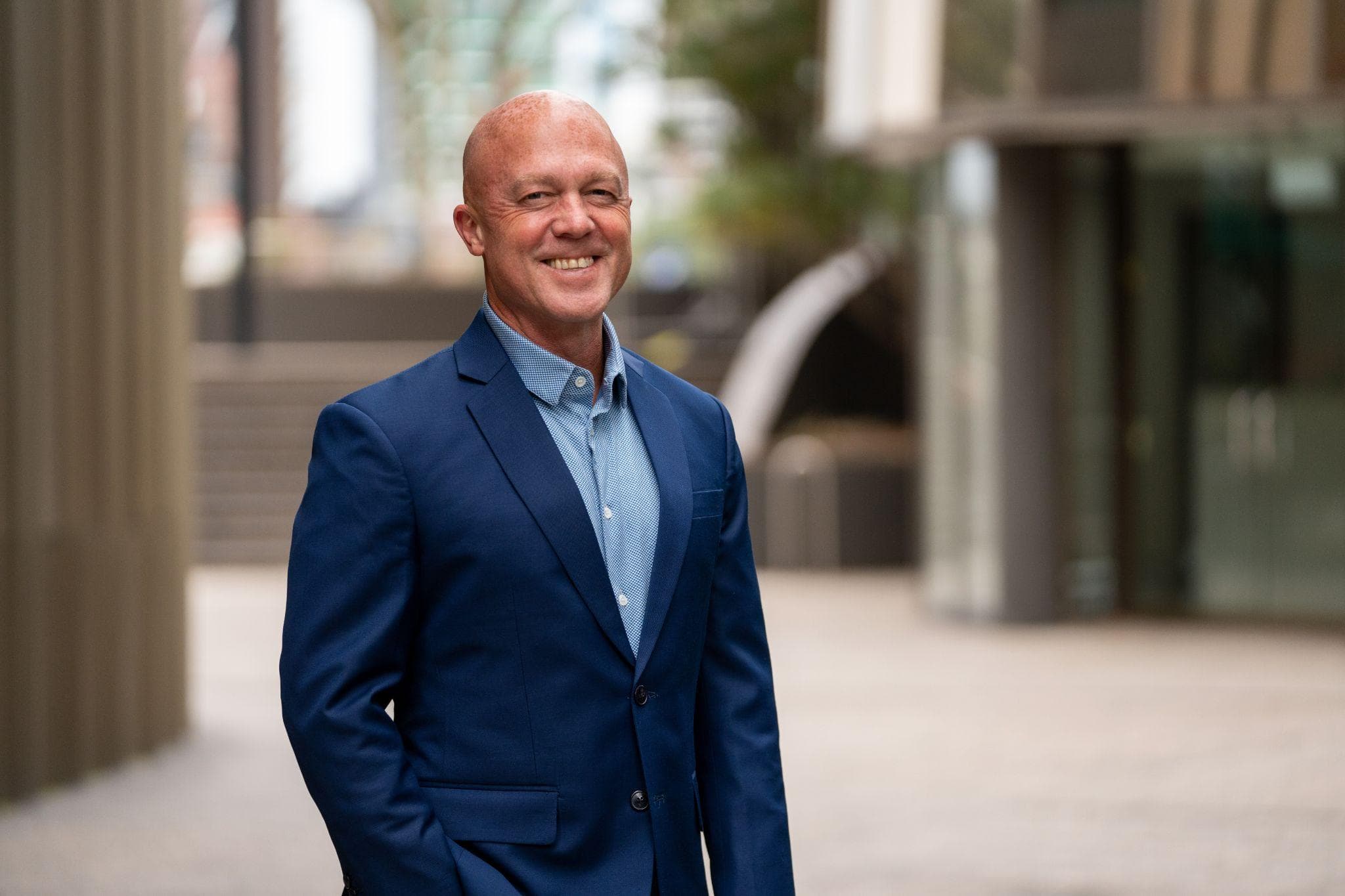 Mark Davis smiling, in a navy blazer and light blue shirt, standing in a city plaza.